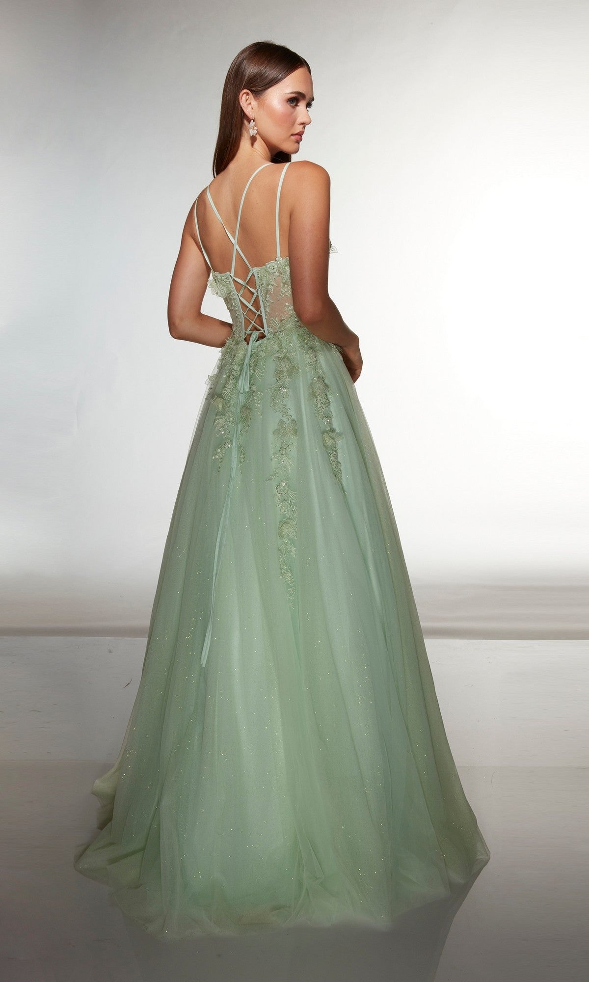 A woman models the Alyce 62203 long prom dress in light green, featuring 3D floral appliqué, thin straps, and a lace-up back, standing against a plain background.